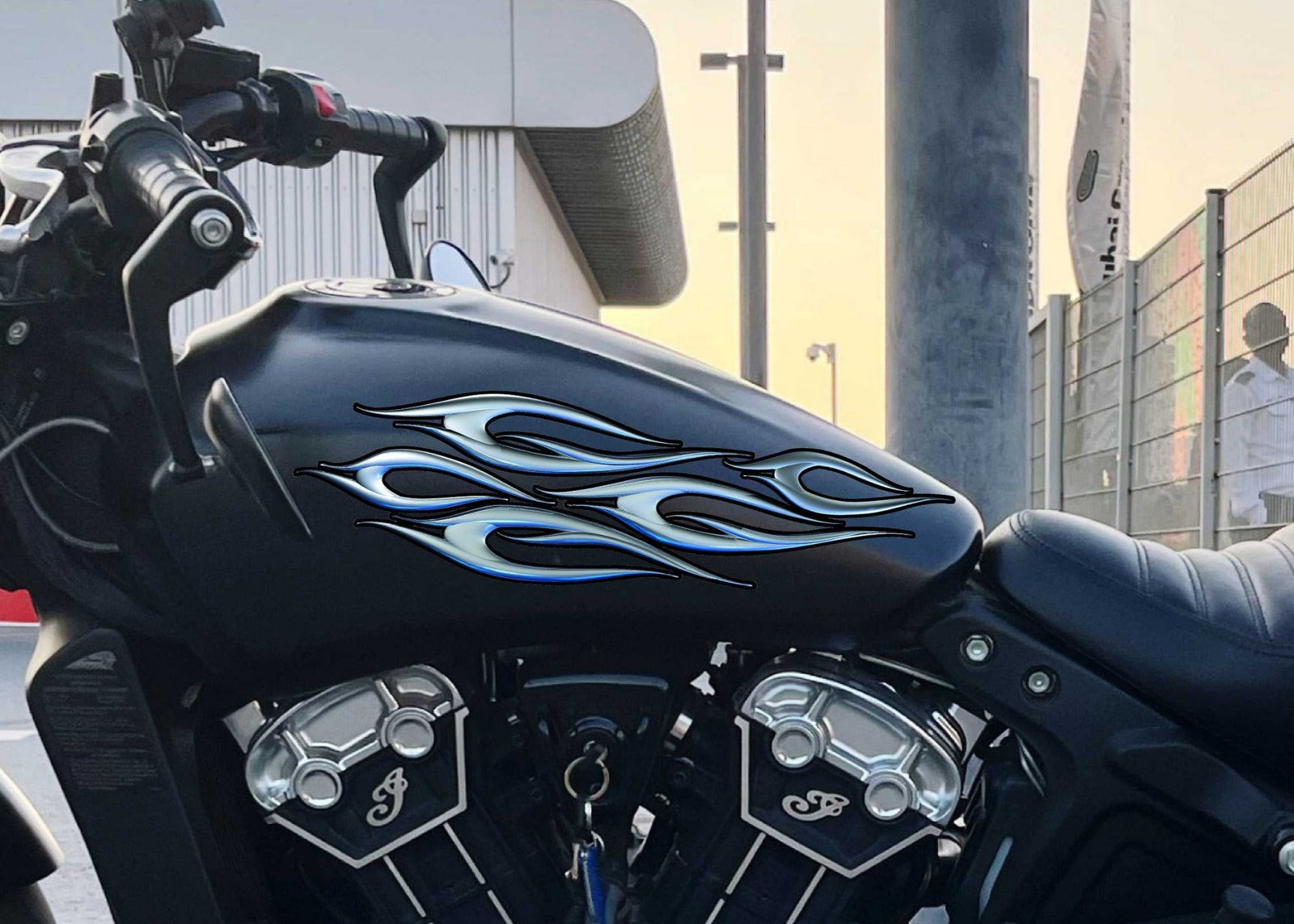 Black motorcycle with bluish chrome decal on its gas tank, parked on a concrete surface with a building and fence in the background.