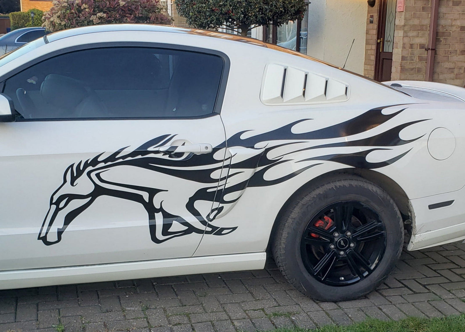 White car with a black mustang horse and flames decal parked on a driveway.
