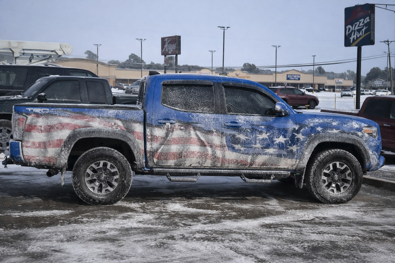 Blue pickup truck with an American flag side wrap covered in winter road salt and slush, leaving a white hazy buildup across the vinyl graphics.