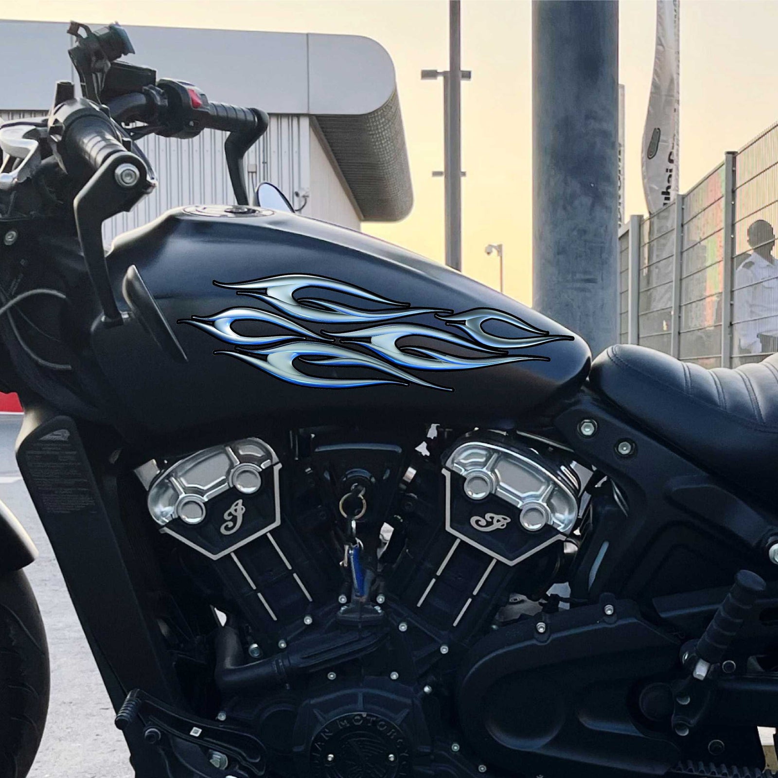 Black motorcycle with bluish chrome decal on its gas tank, parked on a concrete surface with a building and fence in the background.