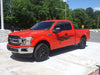 Red pickup truck with black vinyl stripe graphics parked on a paved surface with trees in the background.