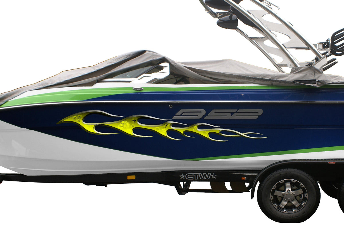 A speedboat on a dual-axle trailer, covered with a dark gray protective cover. The boat features a blue and white hull with green stripes, a yellow skulls flame graphic, and a wakeboard tower on top.